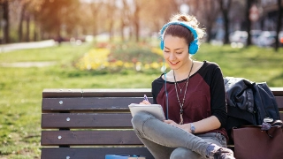 Eine junge Erwachsene sitzt an einem sonnigen Tag auf einer Bank im Park, hört Musik und schreibt Notizen.